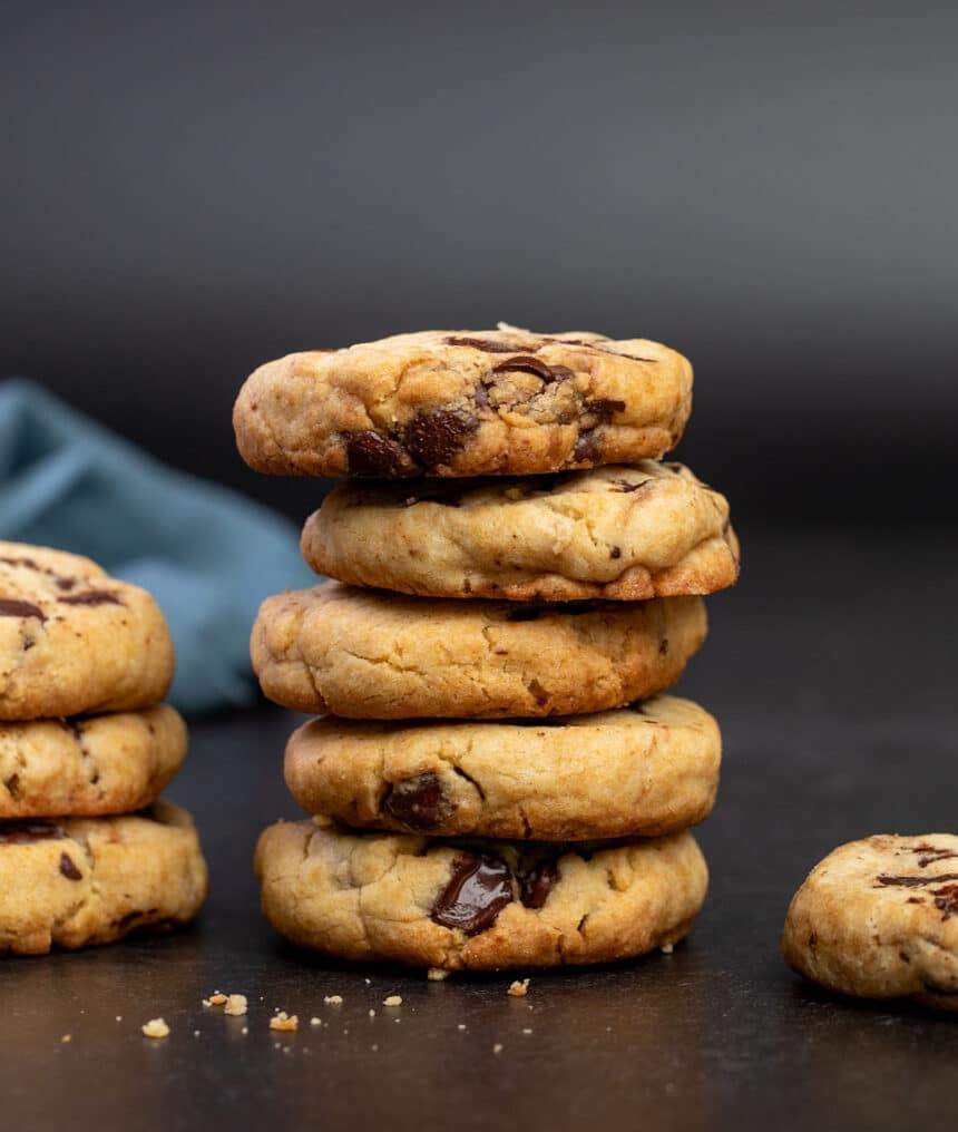 stack of 5 brown cookies with chocolate chips and some crumbs on black surface with blue cloth in background