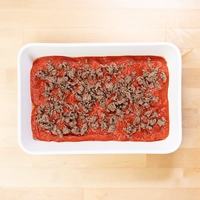 Browned ground beef scattered on top of tomato sauce in white casserole dish