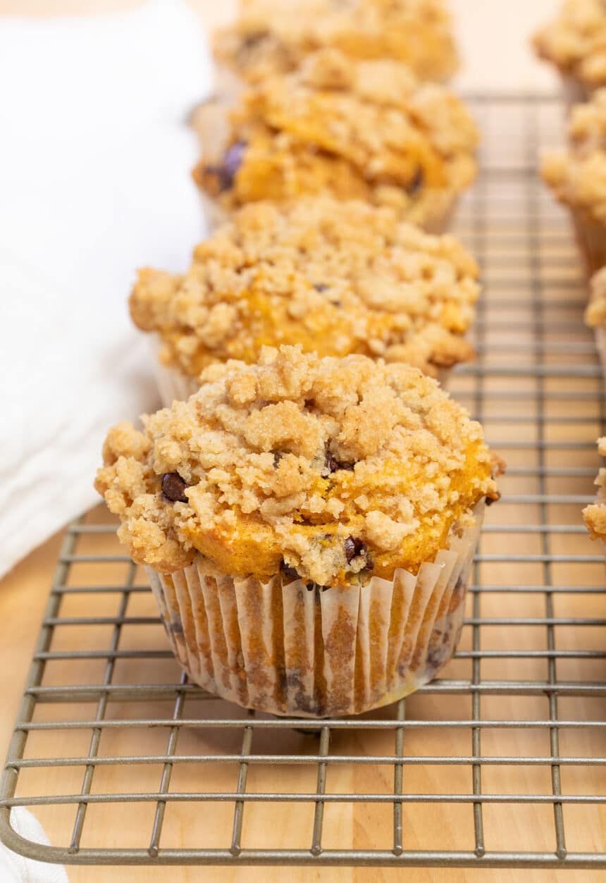 light orange gluten free pumpkin muffins in white liners with crumble topping on wire cooling rack with white cloth