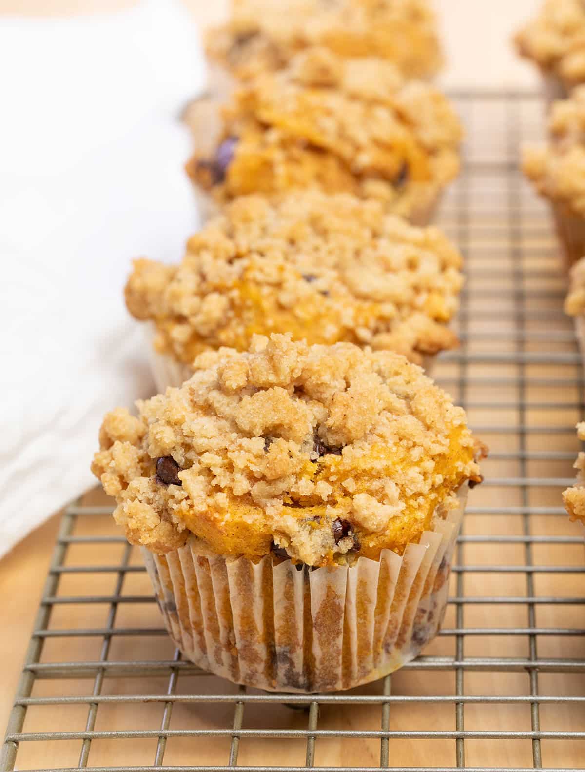 light orange gluten free pumpkin muffins in white liners with crumble topping on wire cooling rack with white cloth