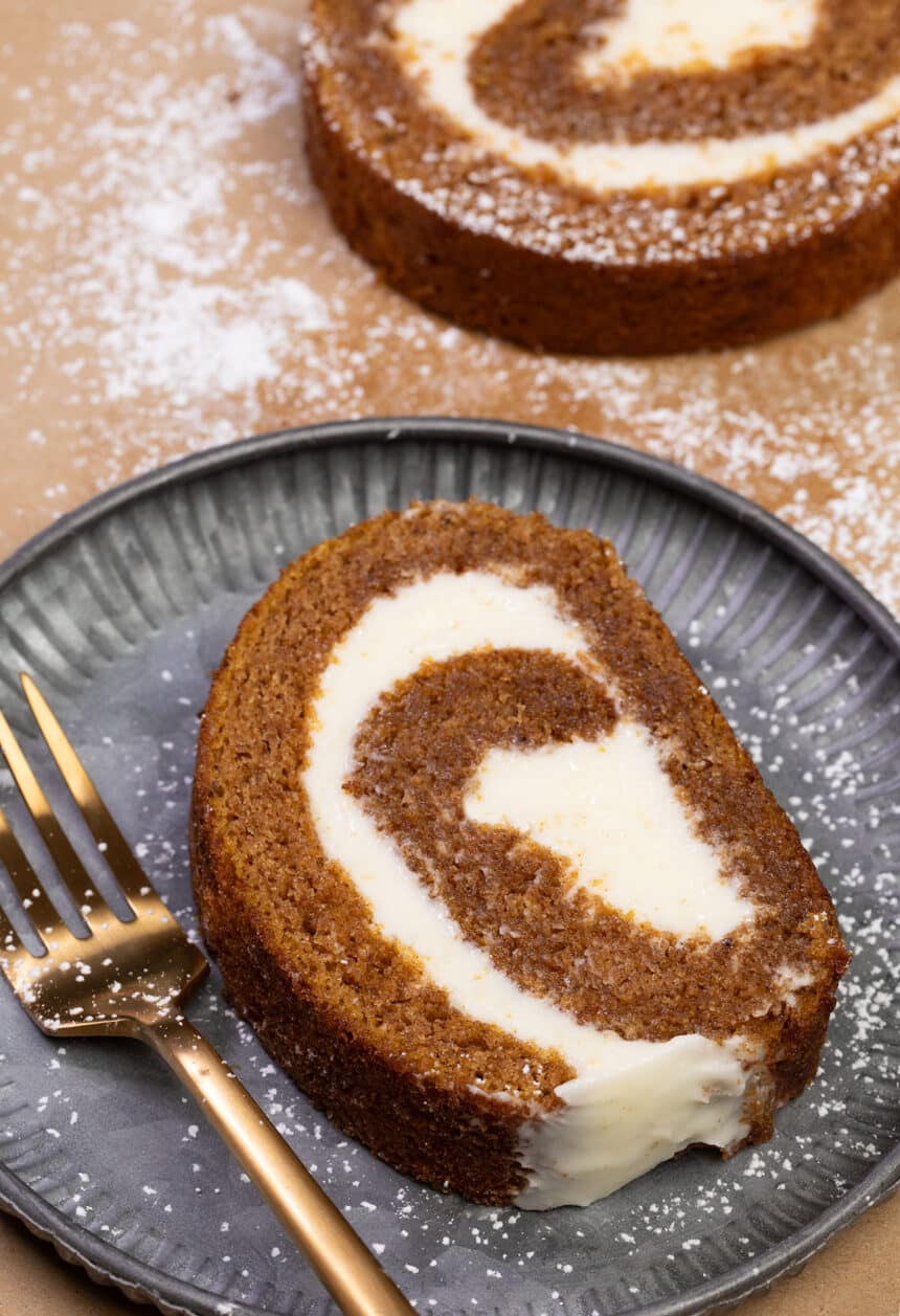 slice of gluten free pumpkin roll cake with white cream cheese filling on small gray plate with gold fork on brown paper