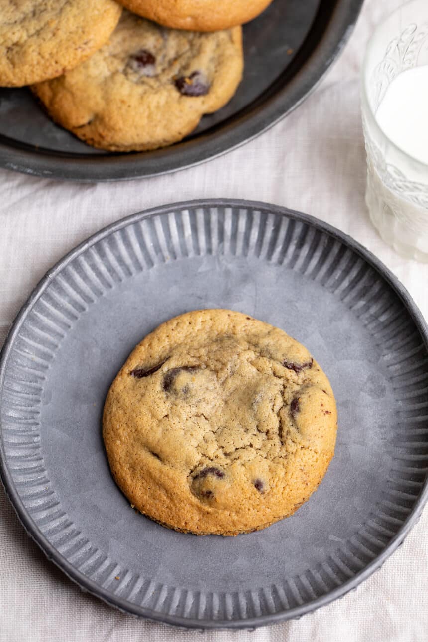 one light brown cookie with chocolate chips on small gray plate with glass of milk and larger plate with more cookies on white cloth