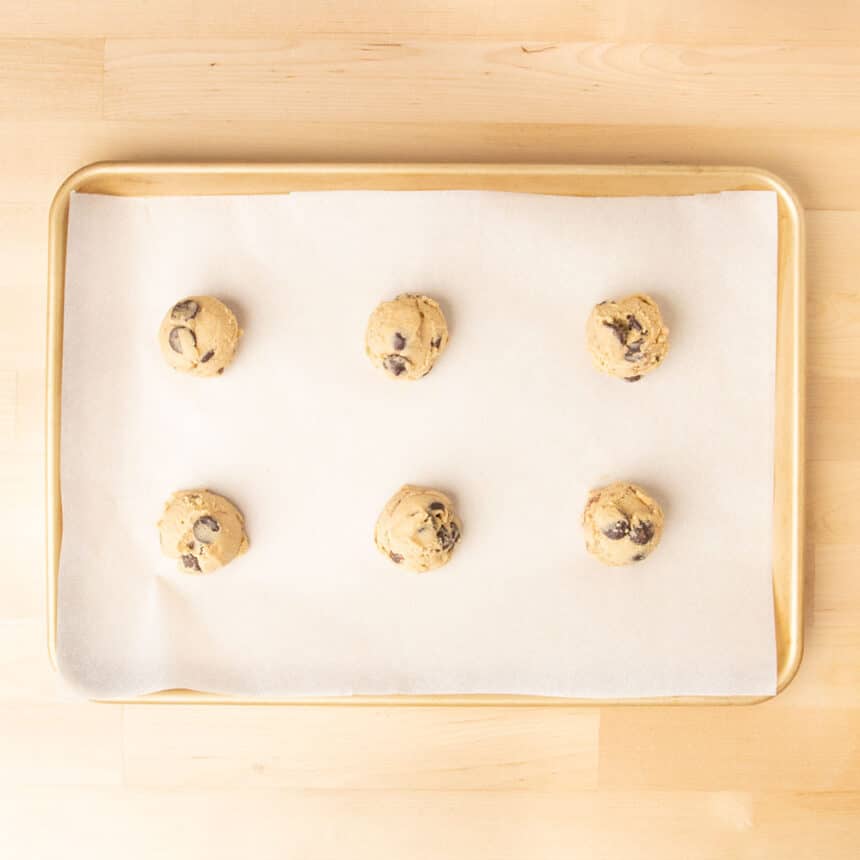 6 balls of raw cookie dough with chocolate chips on paper lined gold baking sheet