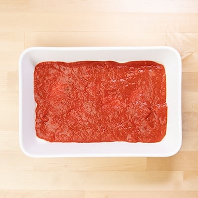 Red tomato sauce on bottom of white casserole dish for gluten free lasagna