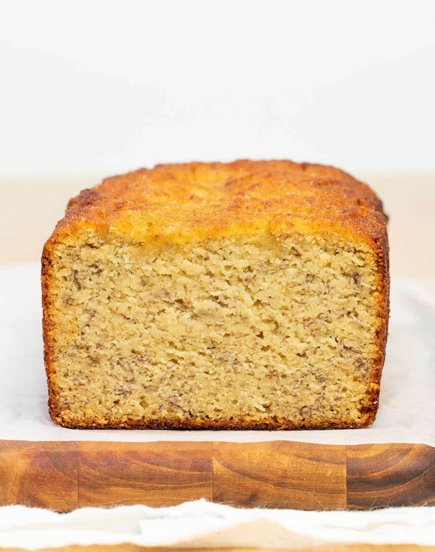 partially sliced loaf of light brown banana bread with brown flecks on white paper on wooden cutting board