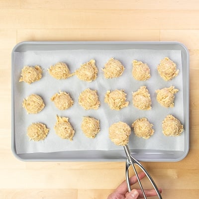 18 raw mounds of chicken meatball mixture with one in hand-held metal ice cream scoop