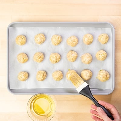 hand holding pastry brush with black handle, brushing yellow olive oil on raw chicken meatballs