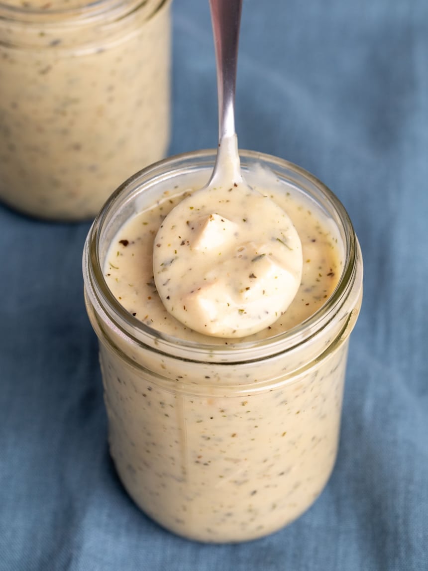 silver soup spoon with light brown gluten free cream of chicken soup with herb flecks and chunks of chicken in glass mason jar on blue cloth