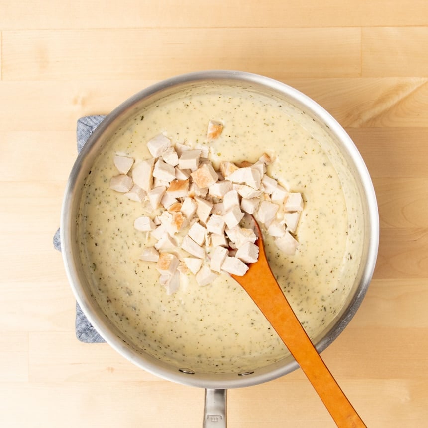 light brown cream soup with herb flecks with brown mixing spoon and cubed cooked chicken in same saucepan