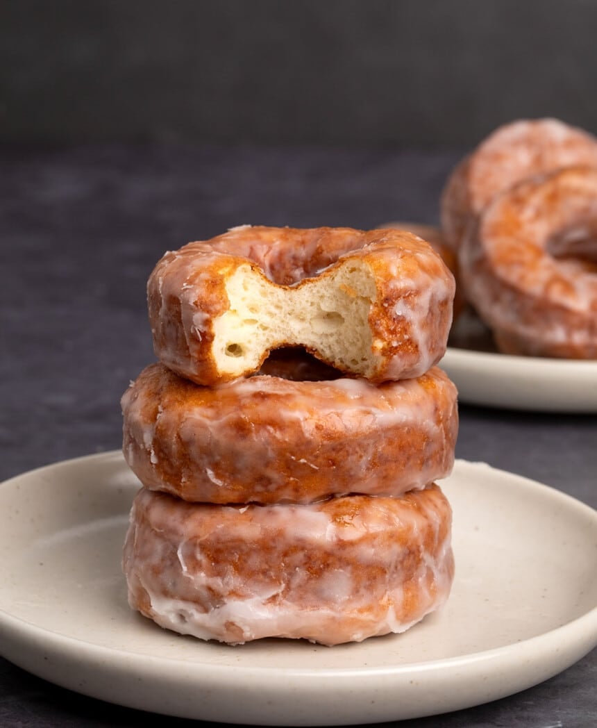 stack of 3 brown gluten free donuts with white glaze on small tan plate with top donut bitten