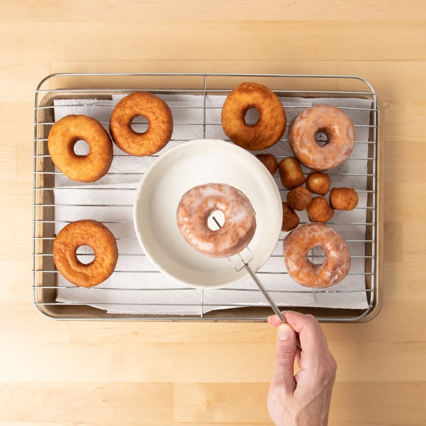 hand holding dipping fork holding brown donut with glaze over wire rack