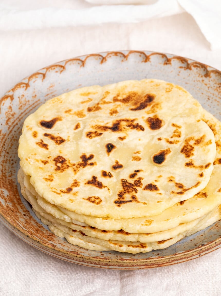 Stack of 6 light brown flatbreads with dark spots in brown round clay plate on white cloth