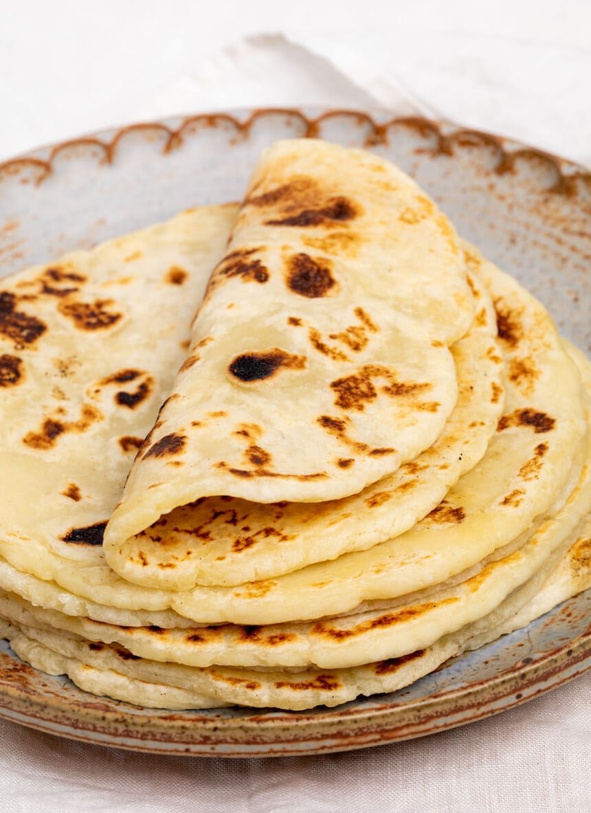 Stack of 8 rounds of light brown flatbread with dark brown spots with top flatbread folded gently in half displaying flexibility of breads