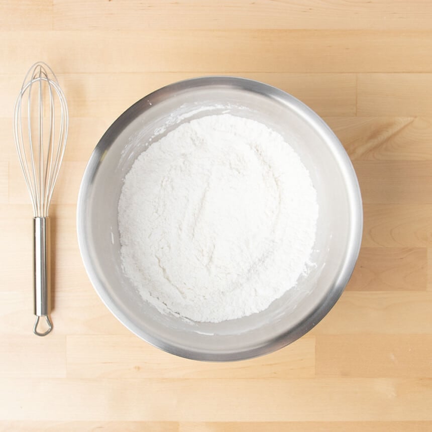 Round metal mixing bowl with white powder and large metal wire whisk on table beside the bowl