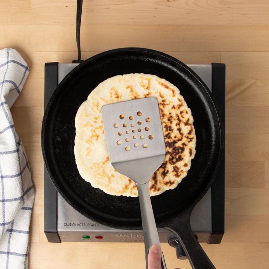 metal spatula pressing on top of round light tan flatbread on black skillet