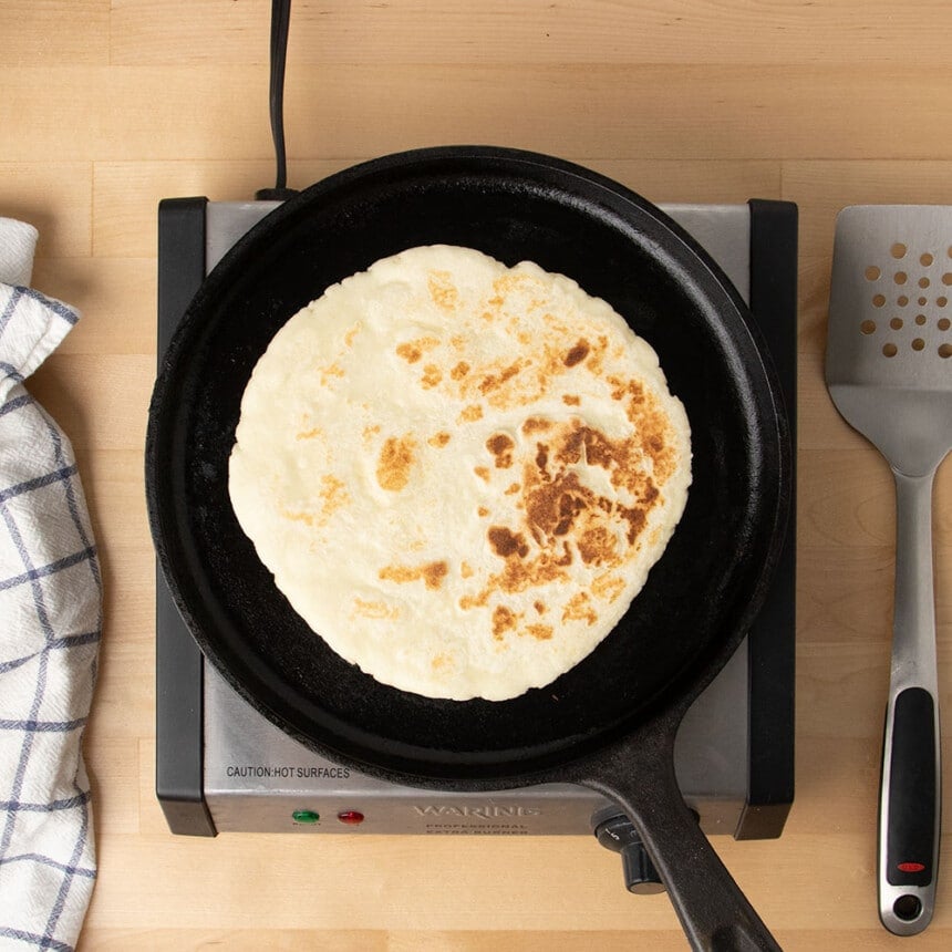 fully cooked light tan round flat bread with brown spots on black cast iron skillet