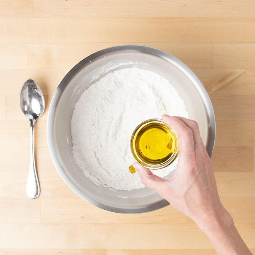 hand holding small round glass bowl with olive oil in it over metal mixing bowl ready to pour oil into the bowl