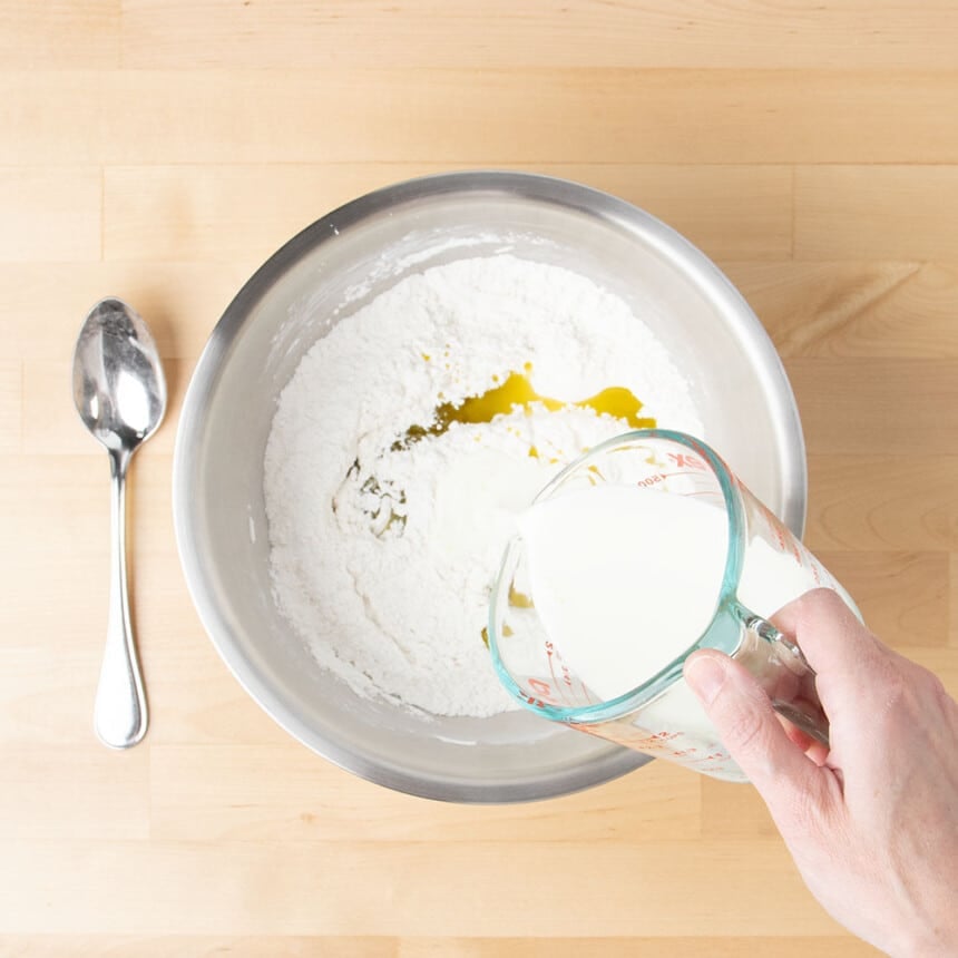 hand holding glass measuring cup pouring milk into same metal mixing bowl