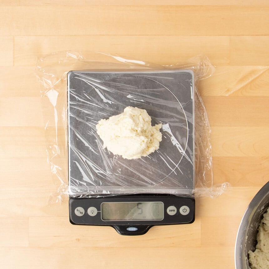 shaggy piece of raw dough on sheet of plastic wrap on metal digital kitchen scale with display reading 5 ounces