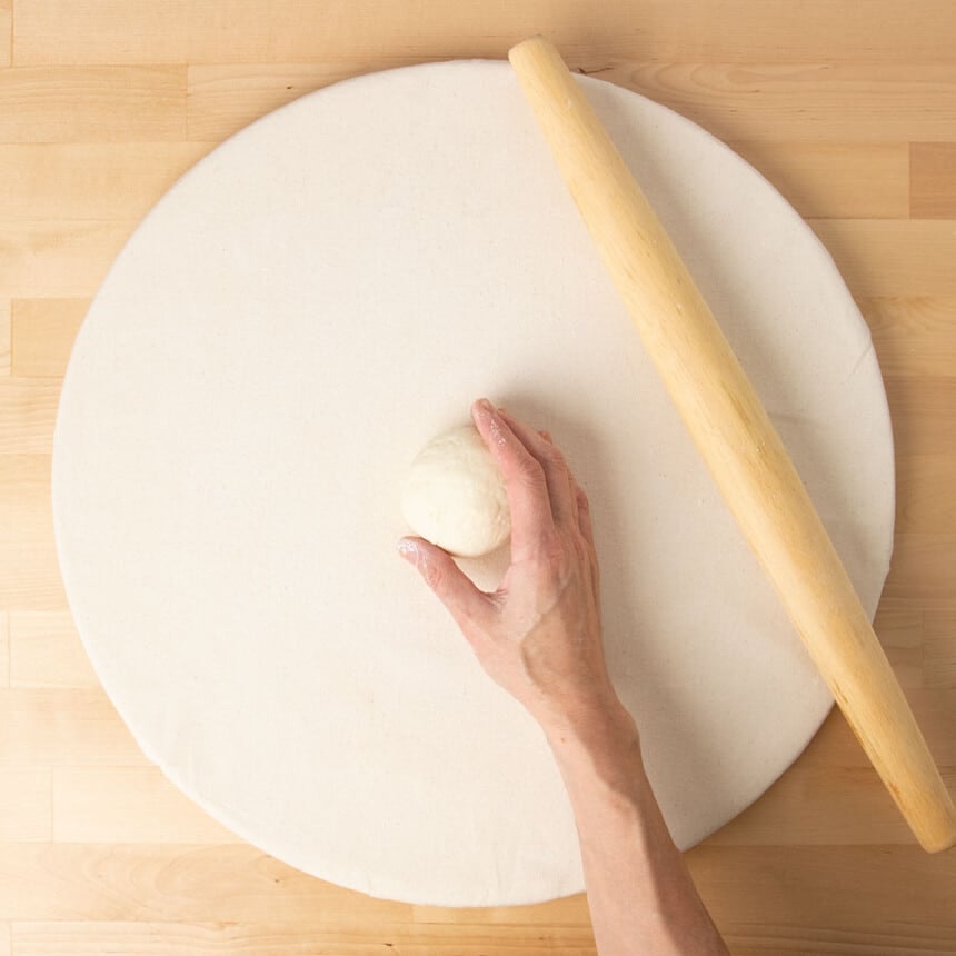 hand cupping sides of round of raw dough to shape it on top of cloth covered round pastry board