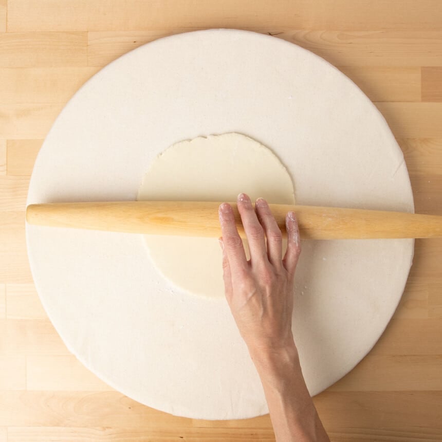 hand rolling French rolling pin over raw dough on cloth covered round board