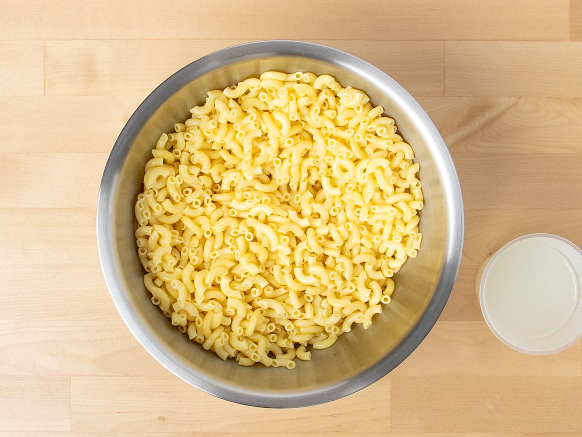 Overhead view of cooked elbow noodles in large metal mixing bowl with cup of pasta water.