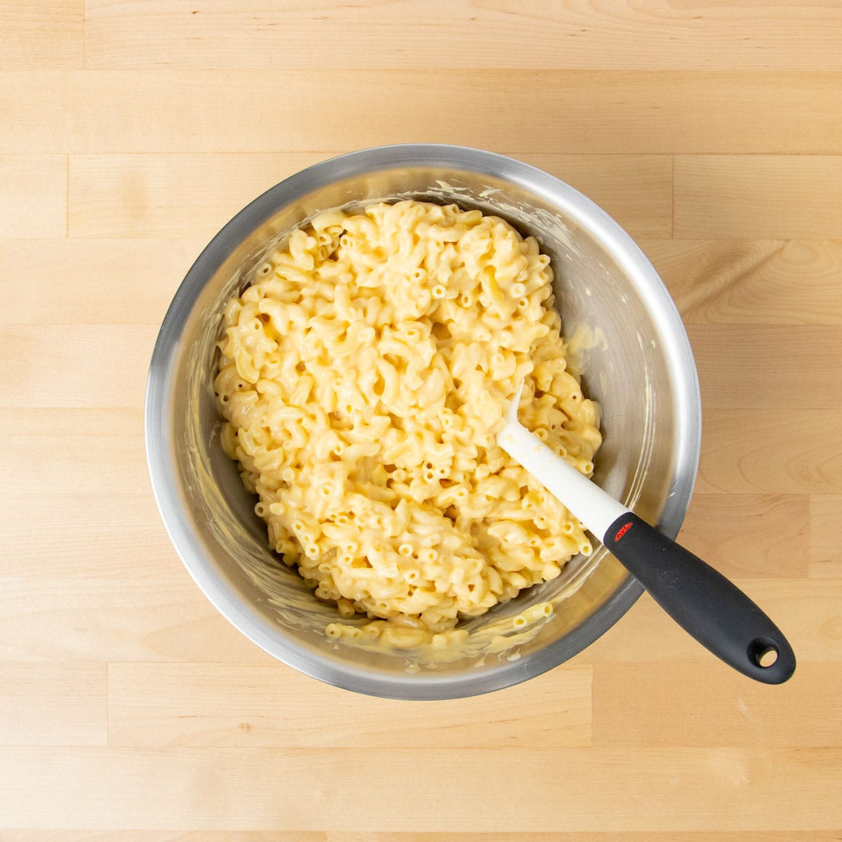 Cooked elbow macaroni mixed with yellow cheese sauce in metal mixing bowl with white spatula with black handle.