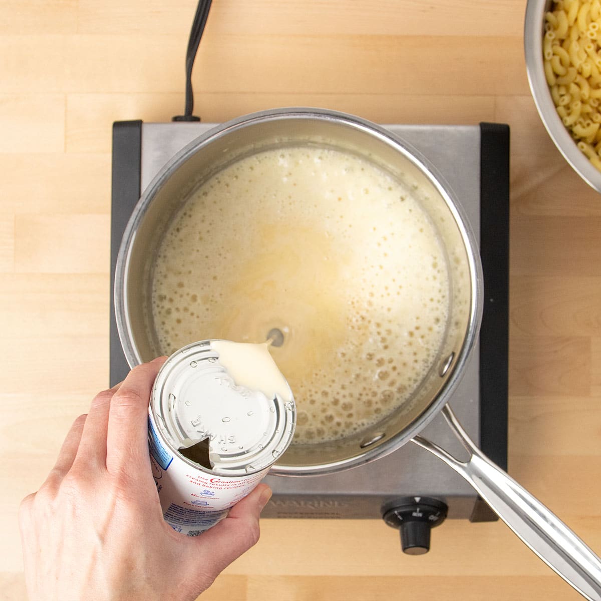 Hand pouring evaporated milk from a metal can into a metal saucepan with light tan bubbling sauce.