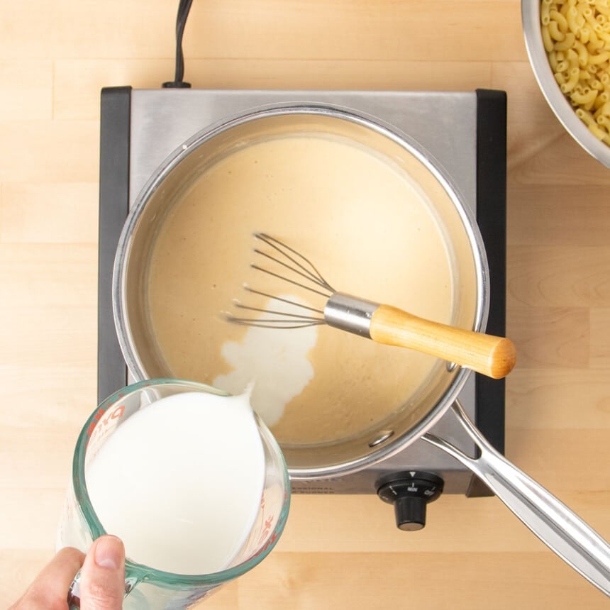 hand pouring milk from glass measuring cup into same saucepan with light tan sauce with flat wire whisk