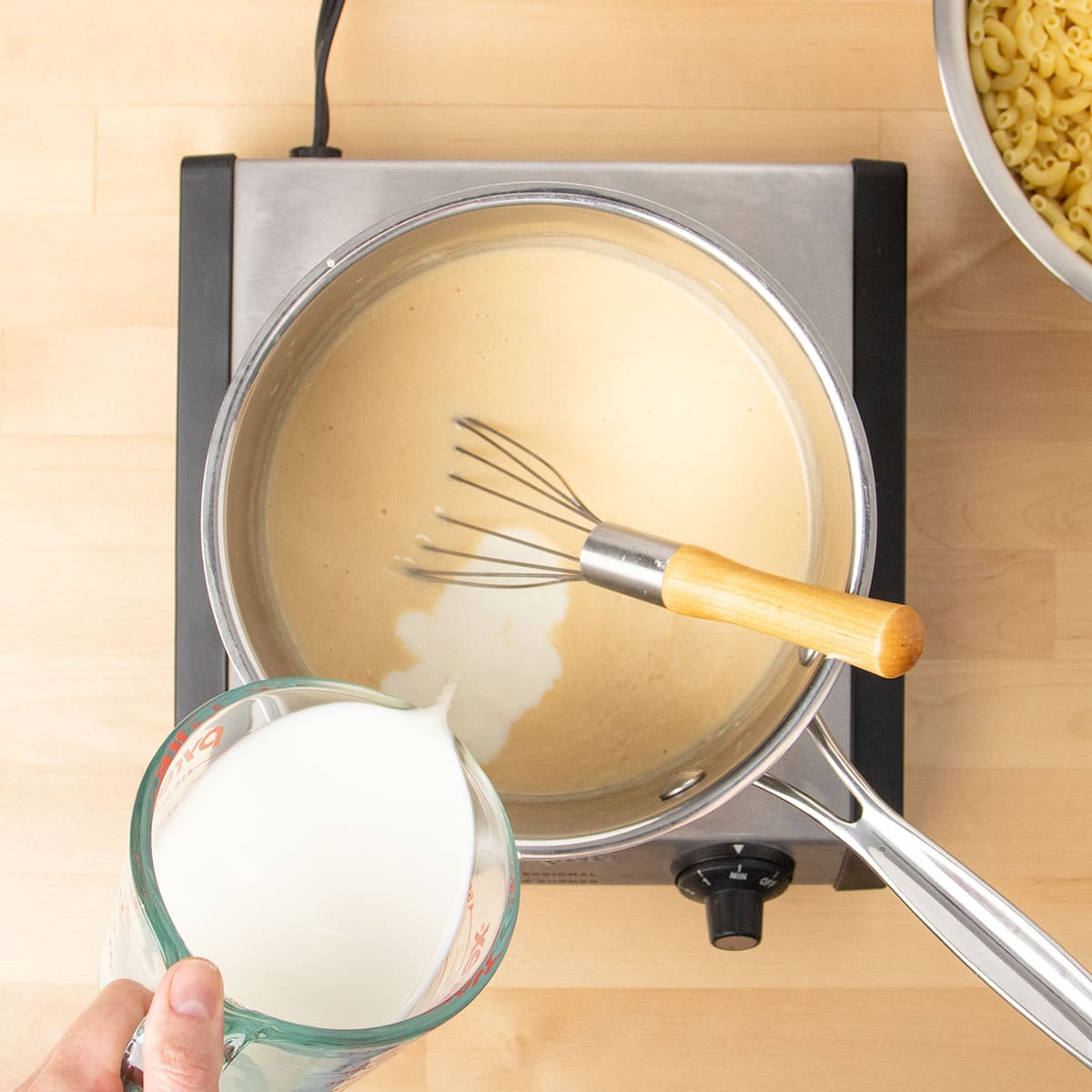 Hand pouring milk from glass measuring cup into same saucepan with light tan sauce with flat wire whisk.