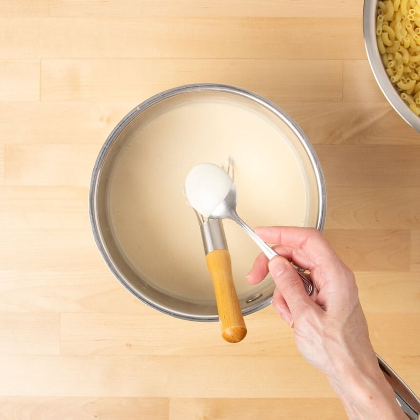 hand holding spoon with light tan sauce coated on back above metal saucepan with rest of sauce