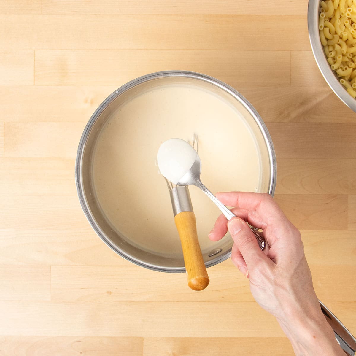 Hand holding spoon with light tan sauce coated on back above metal saucepan with rest of sauce.