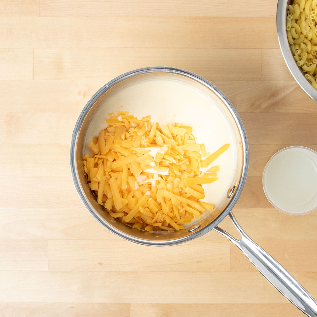 Shredded yellow cheddar cheese in saucepan on top of white sauce.