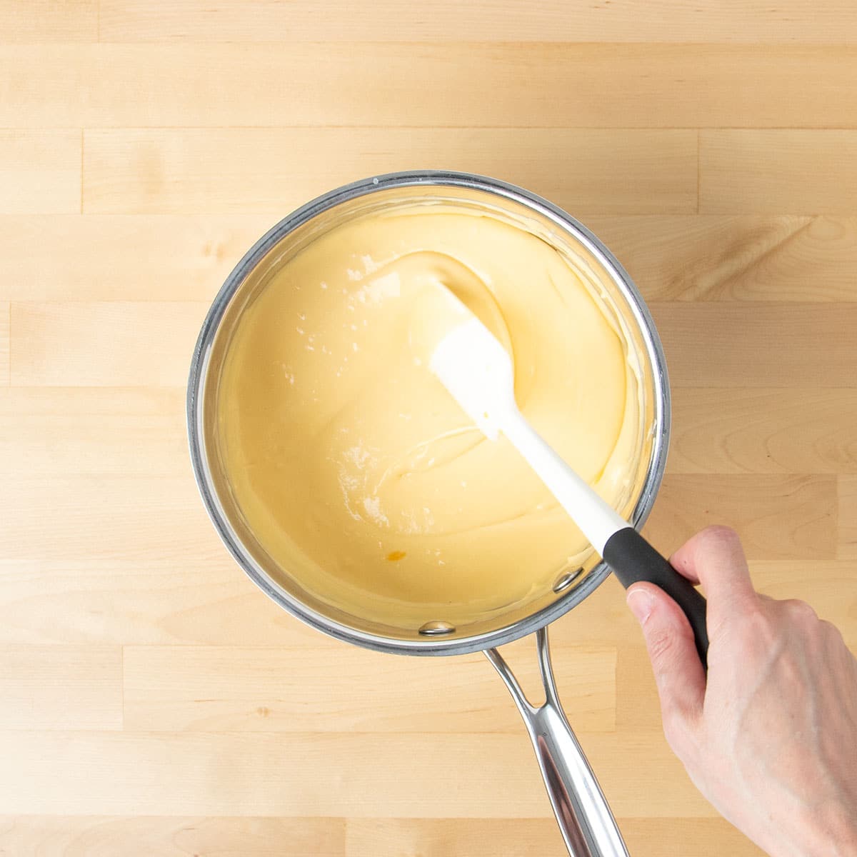 A hand holding white silicone spatula mixing and turning over light yellow cheese sauce in saucepan.