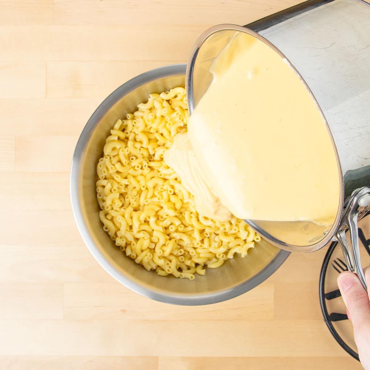 Cheese sauce pouring on top of cooked elbow noodles in metal mixing bowl.