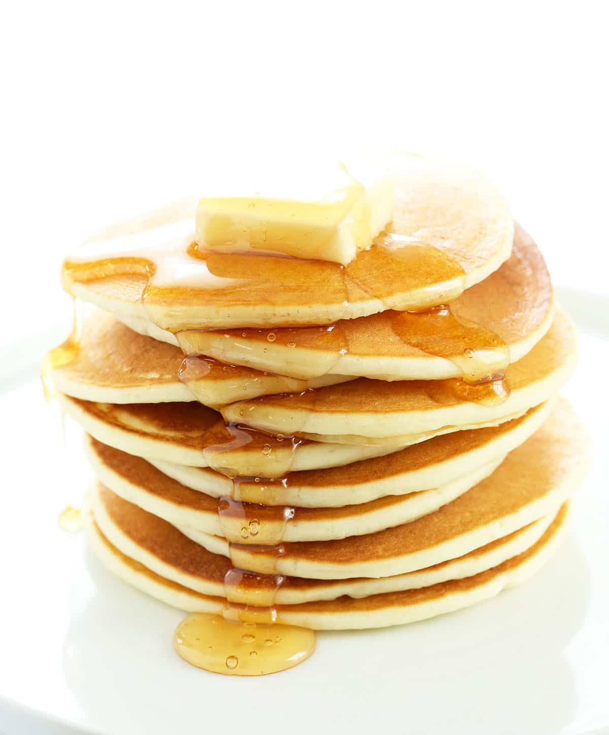stack of 10 golden brown pancakes with pale edges on white plate with pat of yellow butter on top and syrup dripping down the side