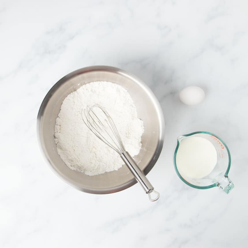 round metal mixing bowl with white dry ingredients and wire whisk with egg and milk in glass measuring cup