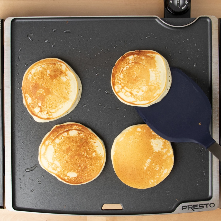 4 light brown cooked pancakes on black griddle with blue silicone spatula picking one up