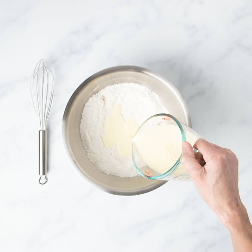 hand pouring egg and milk mixture from glass measuring cup into dry ingredients in same mixing bowl