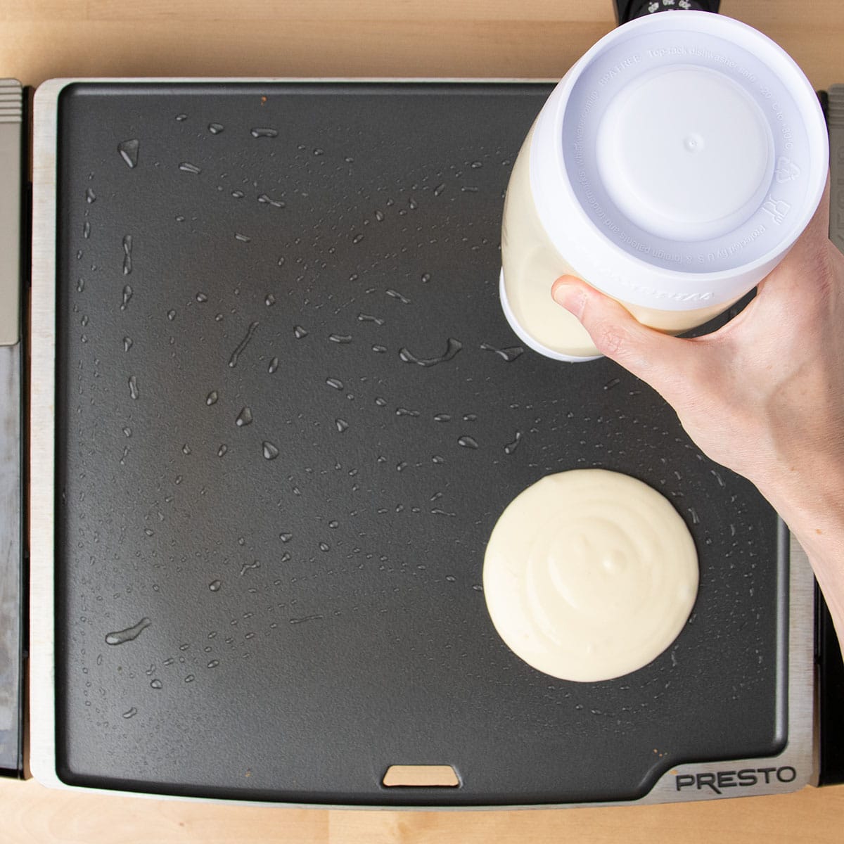 Hand pouring raw pancake mixture onto greased black hot griddle.