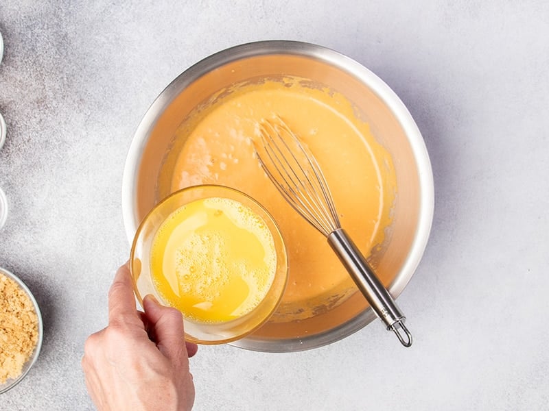 hand pouring yellow mixture from small glass bowl into large round metal mixing bowl with orange liquid and metal whisk