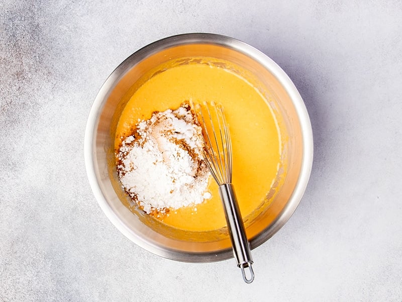 white flour, dark brown pumpkin pie spice sitting on top of orange liquid in same metal mixing bowl with same whisk