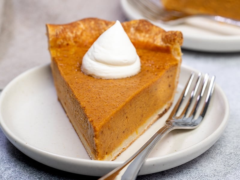 triangular slice of orange pumpkin pie with browned crust and white whipped cream on small cream plate with fork