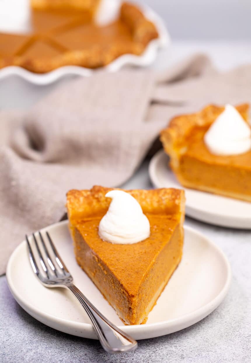 side view of triangular slice of gluten free pumpkin pie on cream plate with fork with tan cloth in background