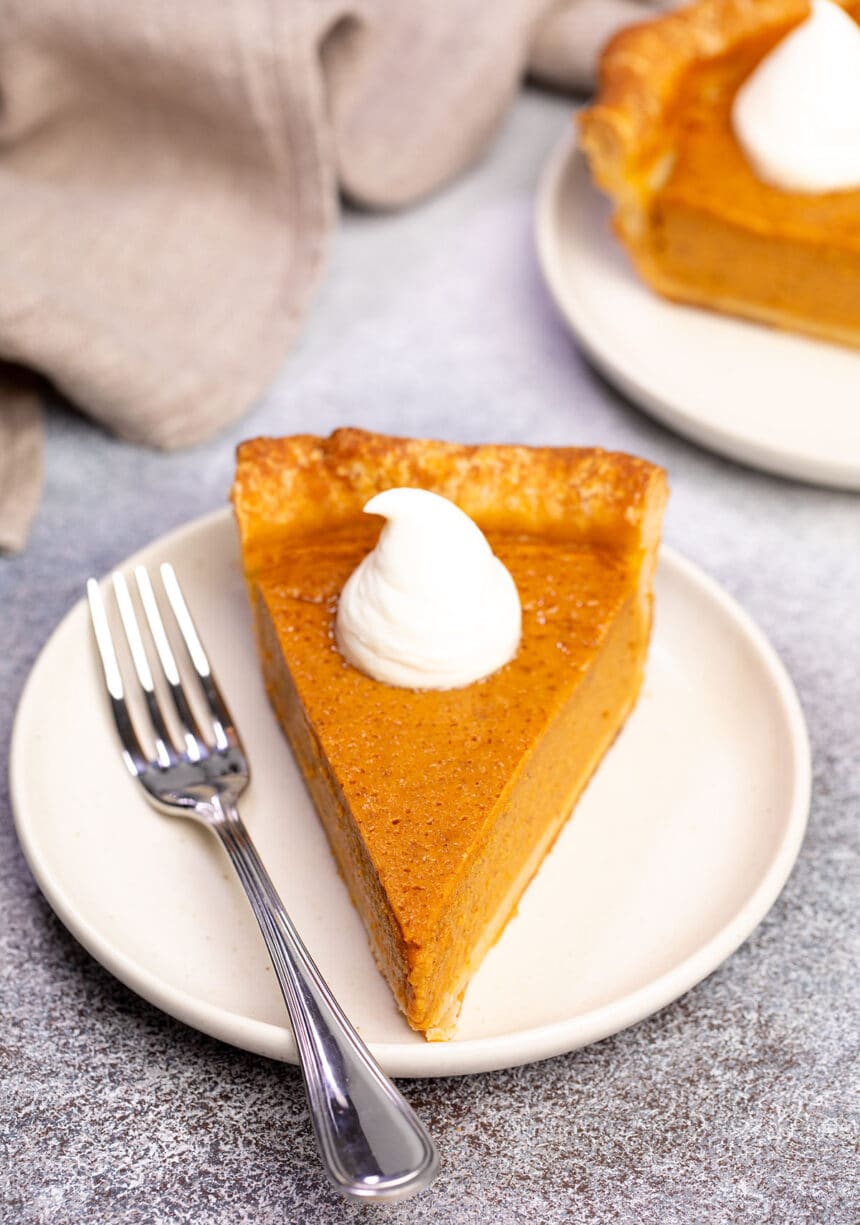 front facing image of triangular slice of orange colored gluten free pumpkin pie with browned pie crust and white dollop of whipped cream on small cream plate with fork