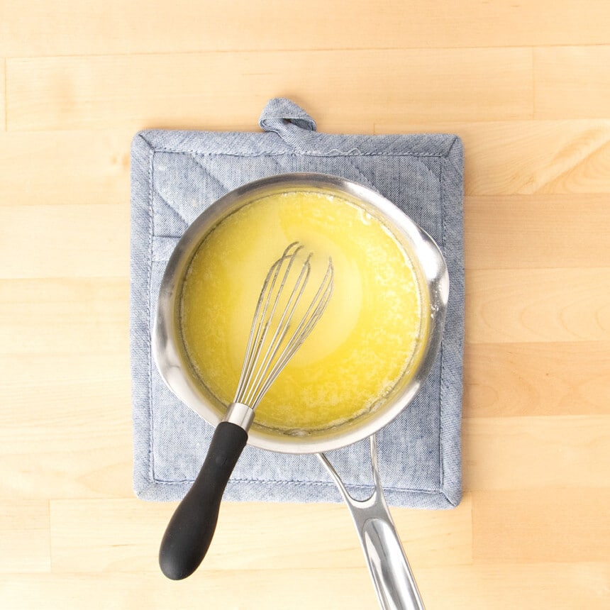 metal whisk with black handle in small metal saucepan with melted butter and sugar on blue pot holder