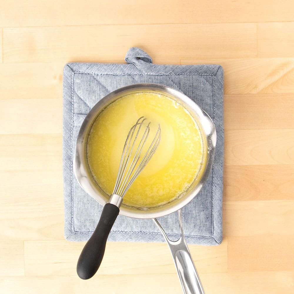 metal whisk with black handle in small metal saucepan with melted butter and sugar on blue pot holder