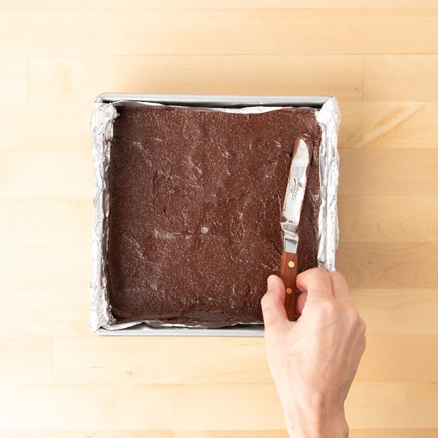 hand holding small offset spatula spreading out dark brown raw brownie batter in metal baking pan