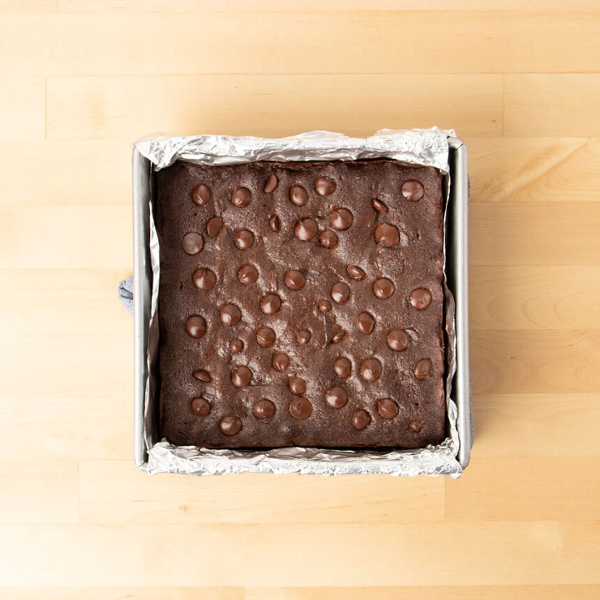 overhead image of uncut baked brownies with chocolate chips in foil-lined square baking pan