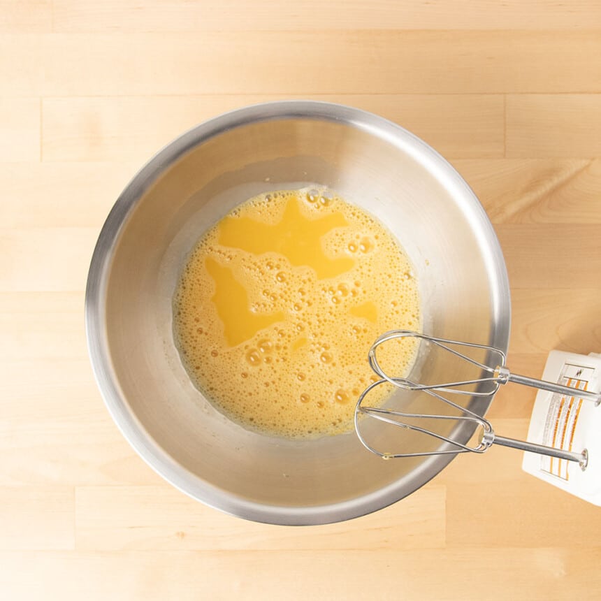 Round metal mixing bowl with frothy beaten eggs and handheld mixer with 2 beaters beside the bowl